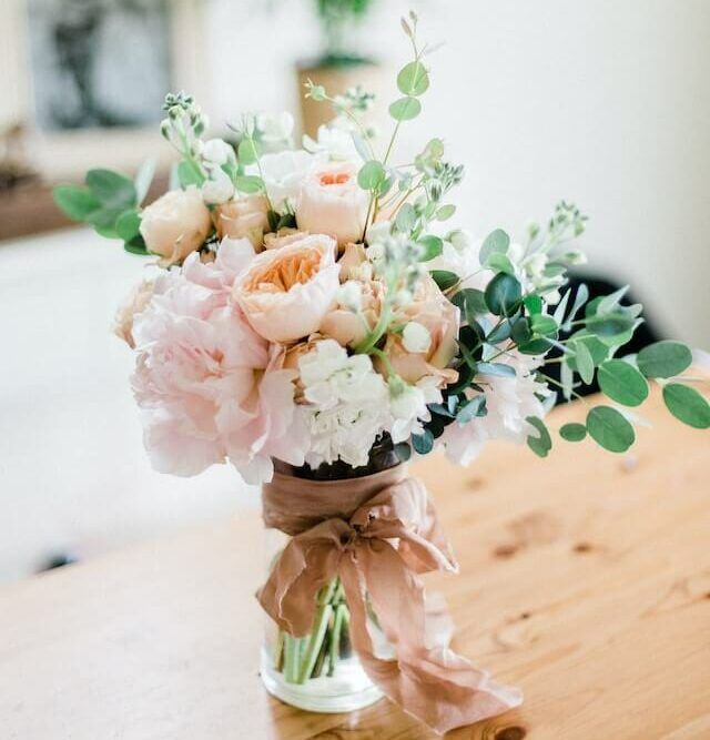 A bouquet of pale pink and peach roses with eucalyptus greenery, tied with a tan ribbon in a glass vase on a wooden table.