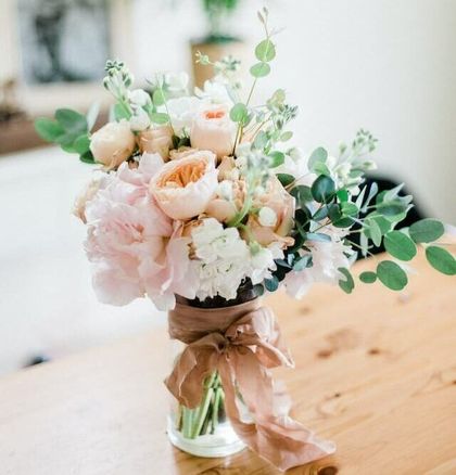 A bouquet of pale pink and peach roses with eucalyptus greenery, tied with a tan ribbon in a glass vase on a wooden table.