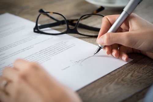 A hand signing a document with a silver pen, with glasses resting on a wooden table in the background.