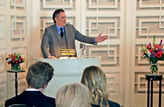 A man in a suit speaks from a podium in a bright room with patterned walls, addressing an audience in front of him.