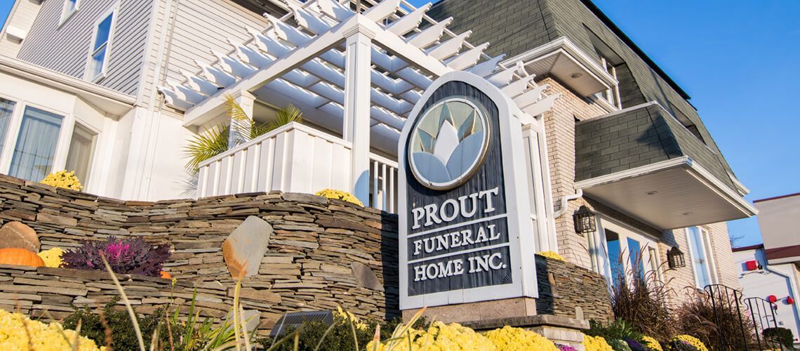 A sign for Prout Funeral Home Inc. stands in front of a white building with a stone retaining wall and yellow flowers.