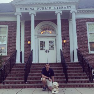 A person kneels with a fluffy dog on the steps of the brick Verona Public Library building.