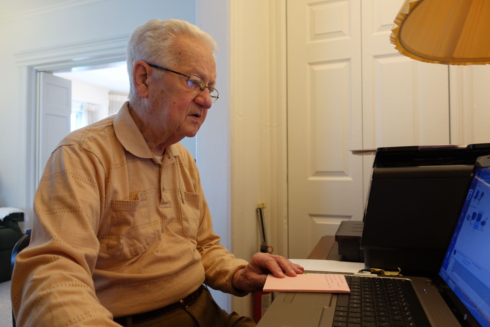 An elderly person in a tan shirt sits at a desk, looking at a laptop screen with a small pink card in their hand.