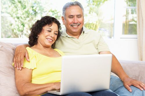 A couple sitting on a couch, smiling while looking at a laptop computer together in a bright, sunlit room.