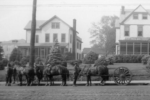A person stands next to a team of four horses pulling a large, spoked-wheel cart in front of two houses on a paved street.