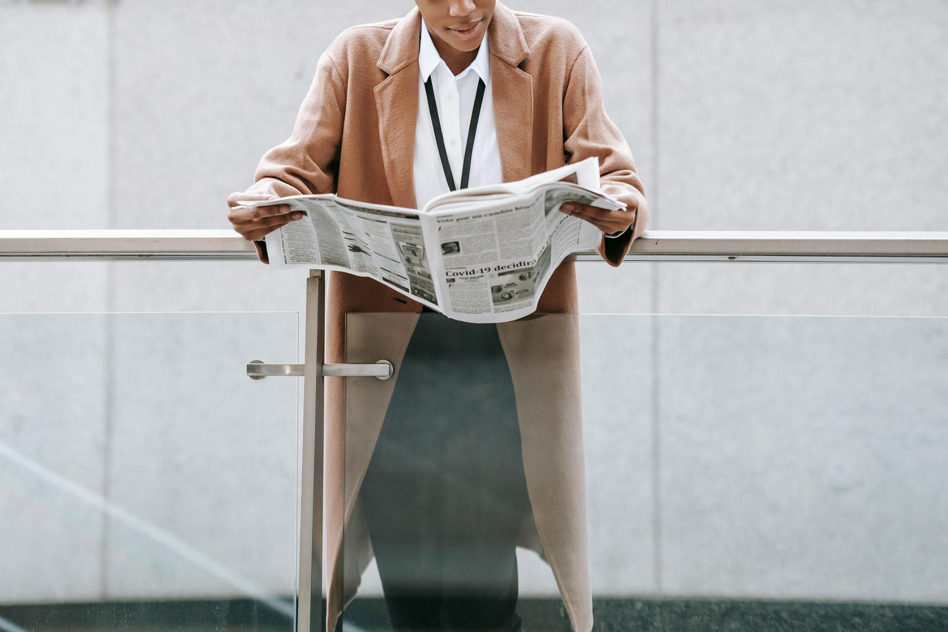 A person in a tan coat and white shirt reads a newspaper while standing behind a glass railing in an office setting.
