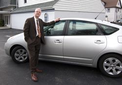 A person in a brown suit stands on an asphalt lot beside a silver sedan, resting a hand on the roof.