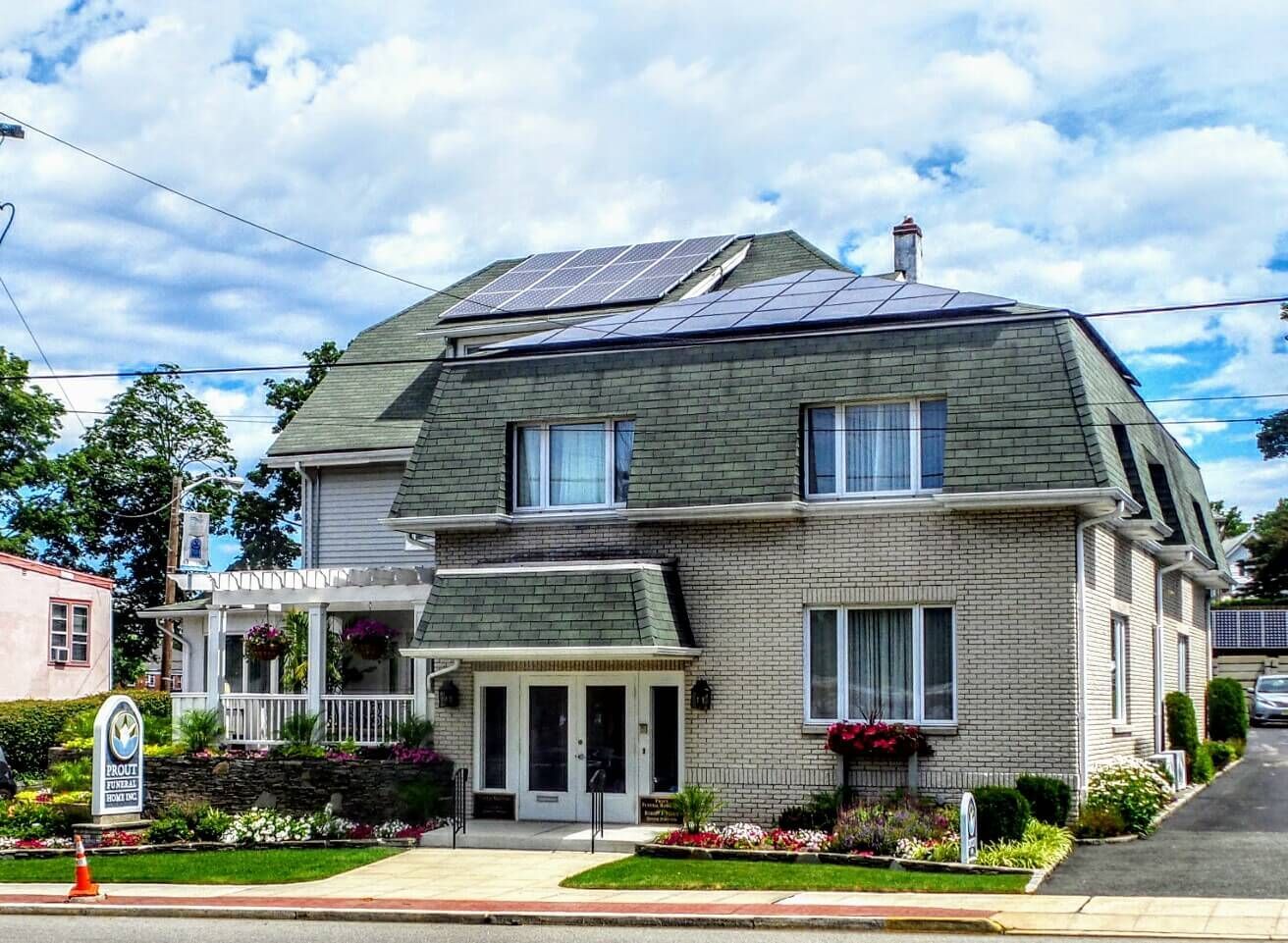 A light-colored two-story building with a dark shingled roof, solar panels, and a front porch under a partly cloudy sky.