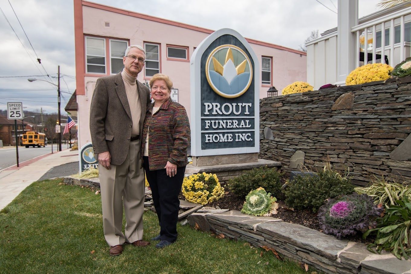 A couple stands next to a sign for Prout Funeral Home Inc. in front of a stone wall and a pale pink building.