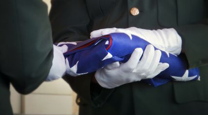 Two service members in dress uniforms and white gloves carefully hold a folded American flag during a ceremony.