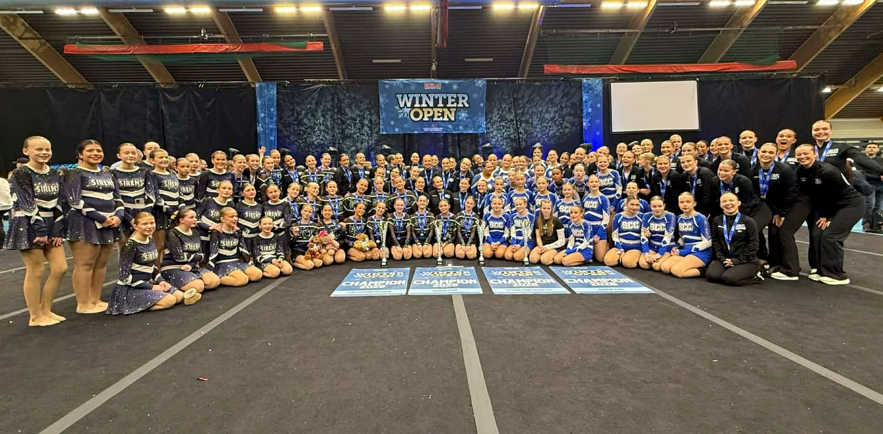 Large group of cheerleaders posing together, some kneeling, on a competition floor, banner in background.