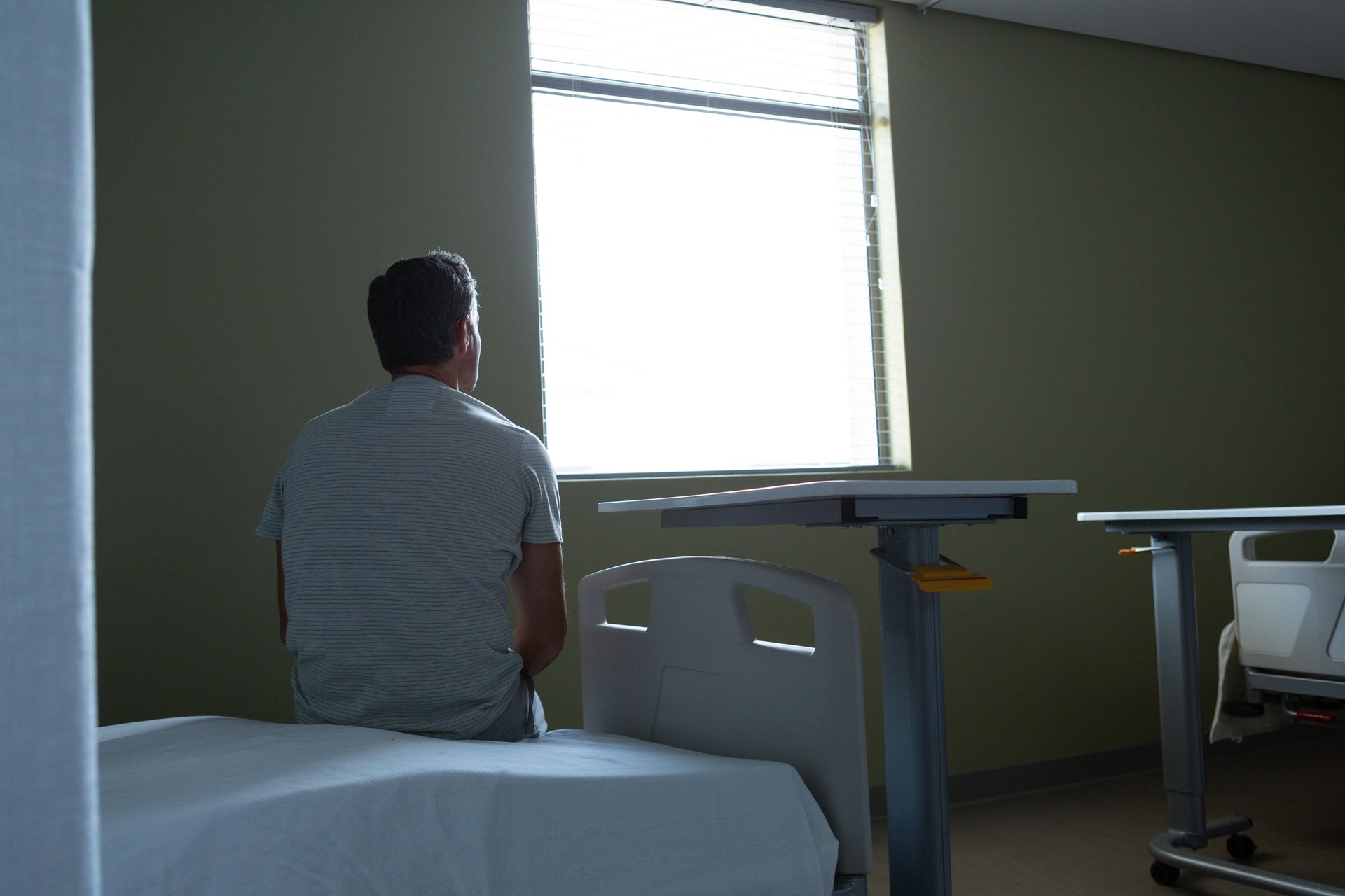 A man is sitting on a hospital bed looking out a window.