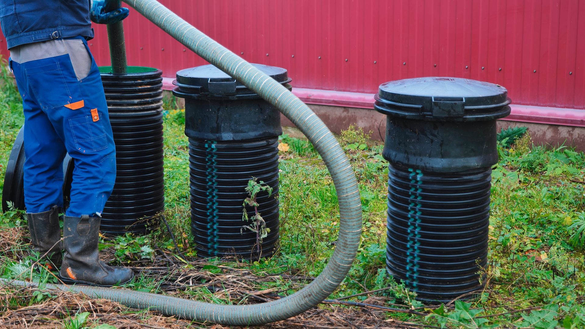 Worker beside three black drainage pipes on wet ground near a red building