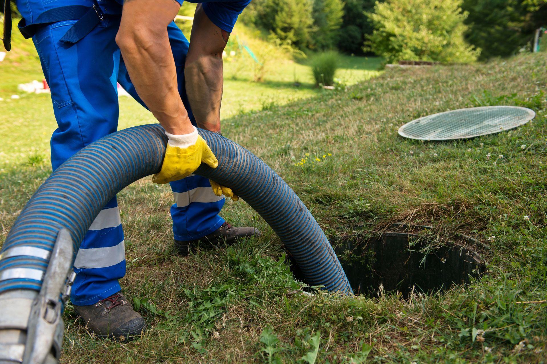 Worker in blue coveralls and yellow gloves handling a large hose at a grassy drainage opening
