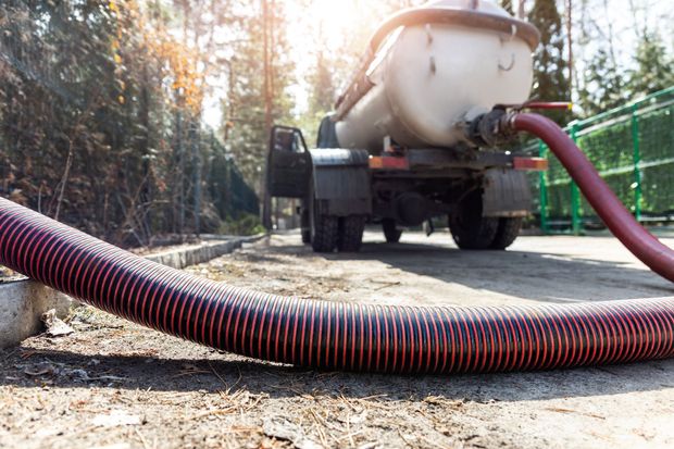 Vacuum truck emptying a red hose onto a dirt road in a wooded area