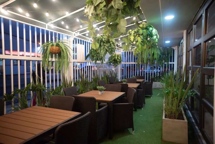 Área de comedor al aire libre con mesas, sillas de mimbre, plantas colgantes y luces de cadena sobre un piso de césped verde.