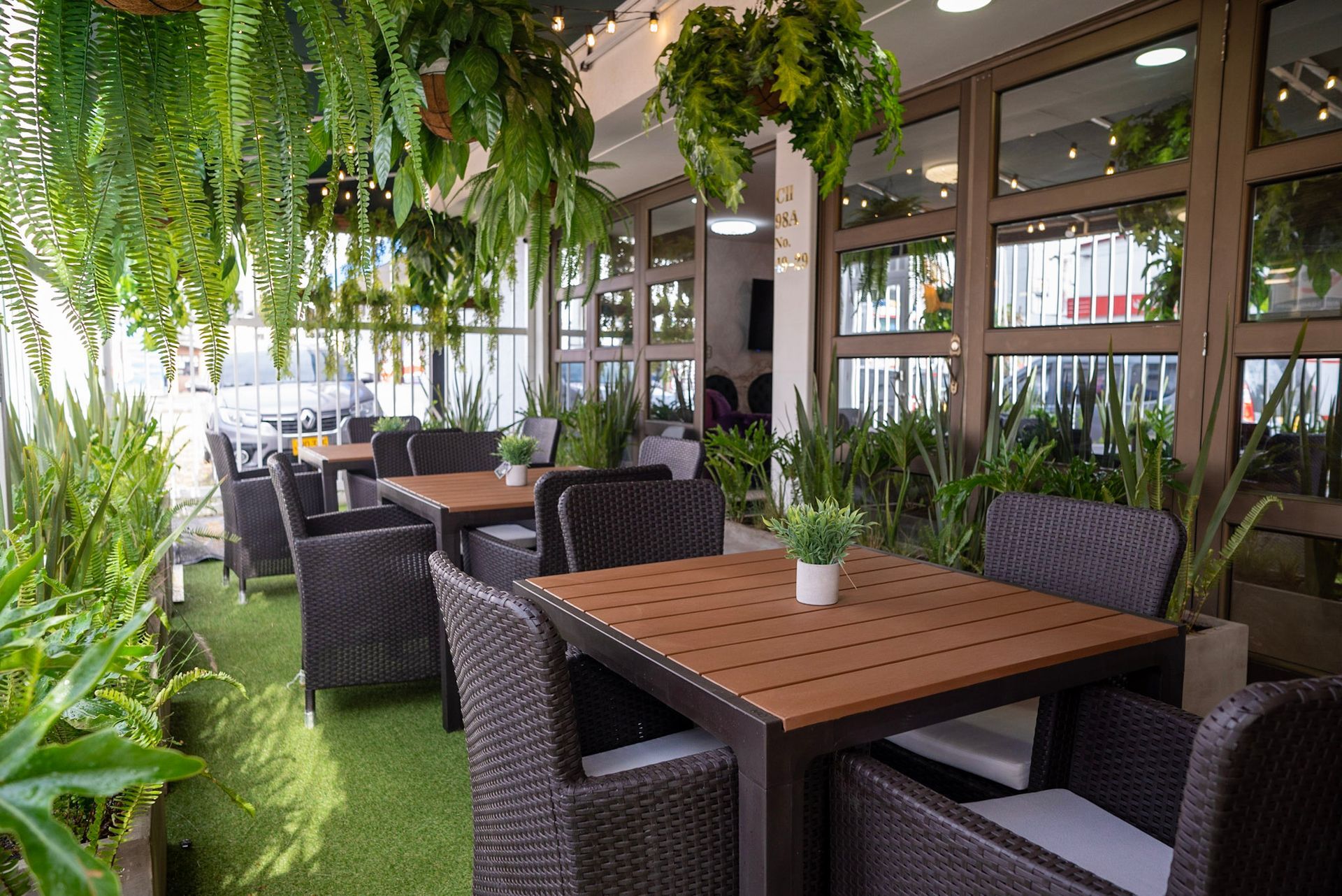 Una zona de estar en el patio con mesas de madera, sillas de mimbre oscuro, césped artificial y plantas colgantes junto a grandes ventanales.