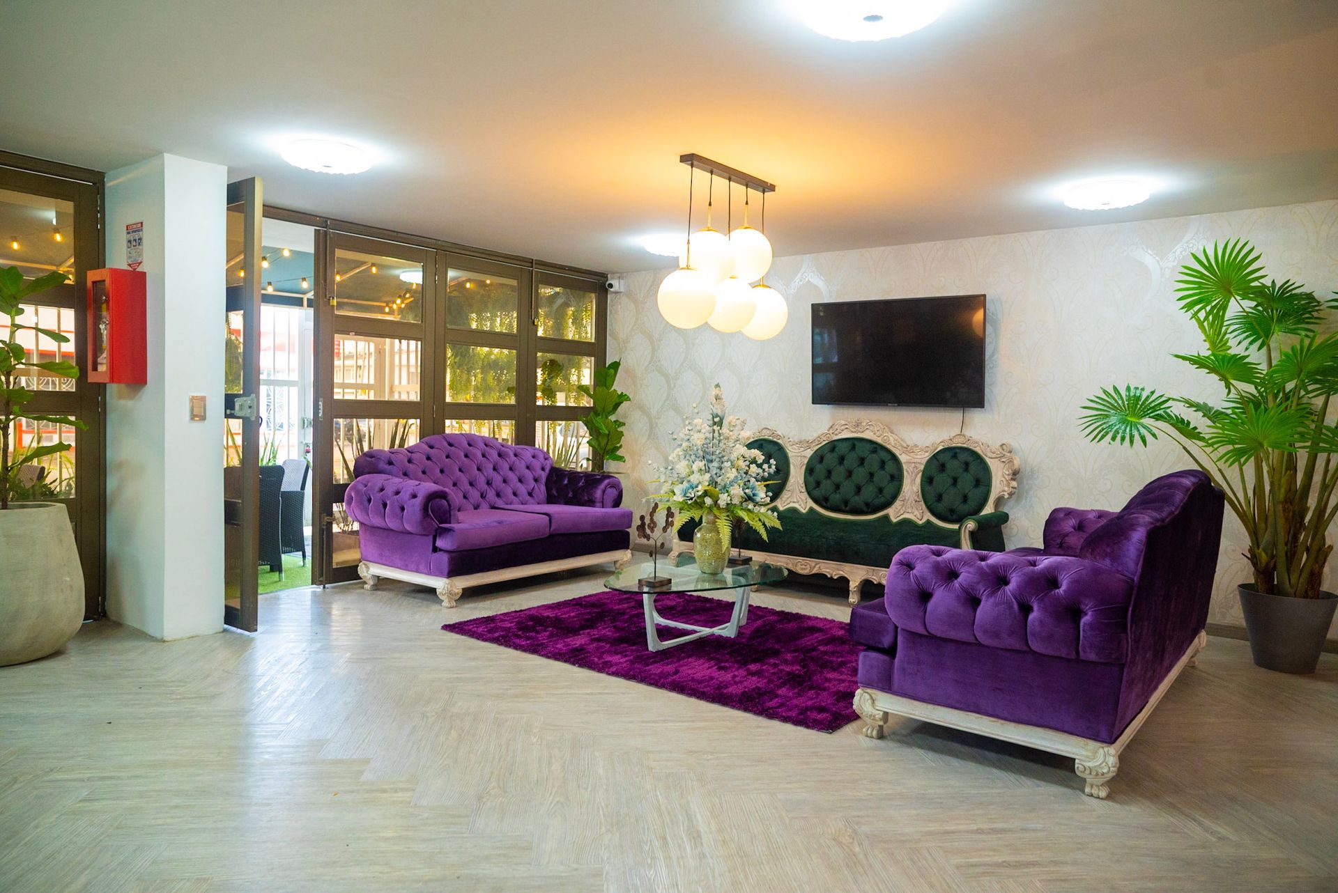 Una sala de estar con dos sofás capitoné de color morado, un sofá central verde, una alfombra morada, una mesa de centro de cristal y grandes plantas de interior.