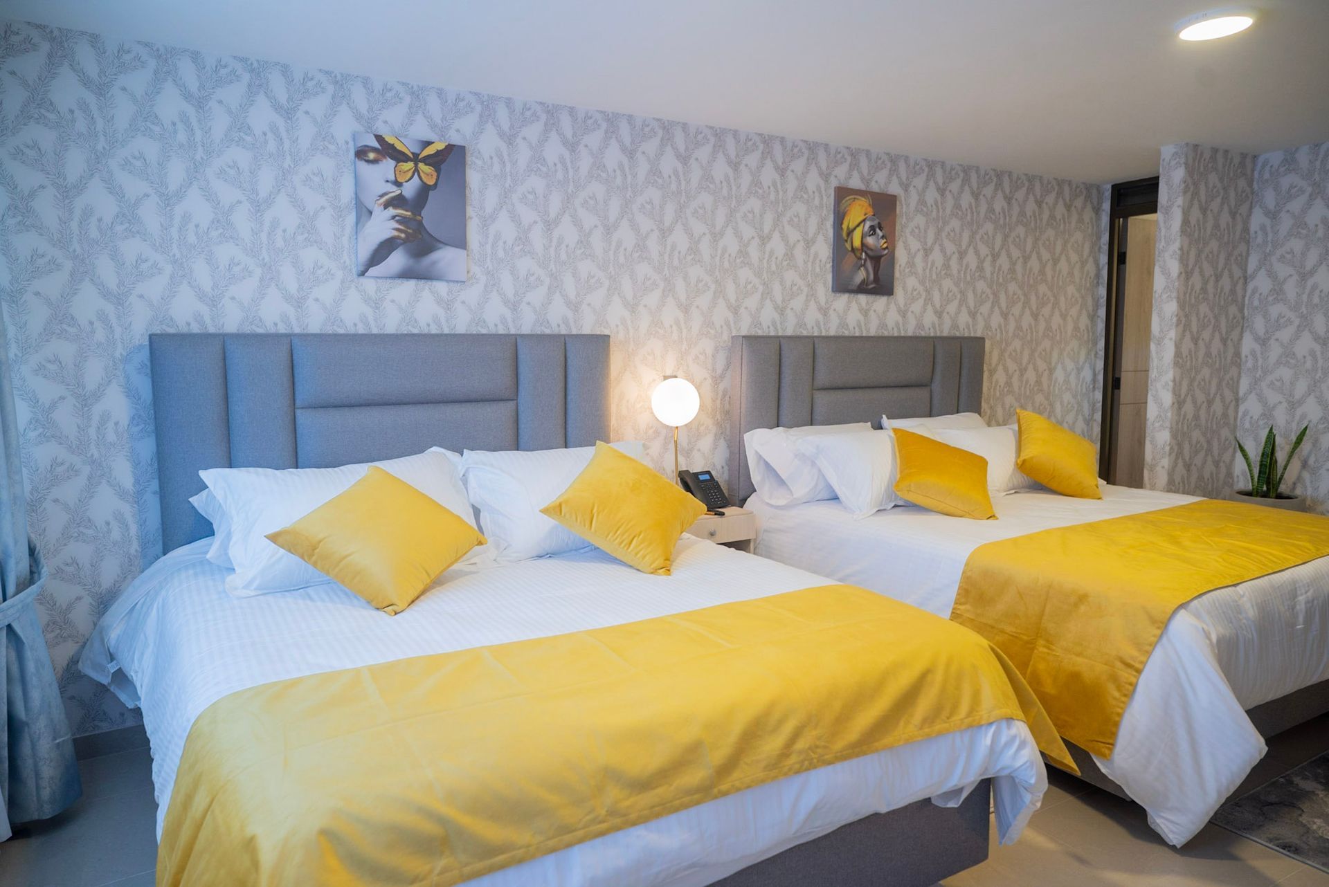 Una habitación de hotel con dos camas tamaño queen, con cabeceras grises, ropa de cama blanca y almohadas y caminos de mesa con detalles en amarillo.