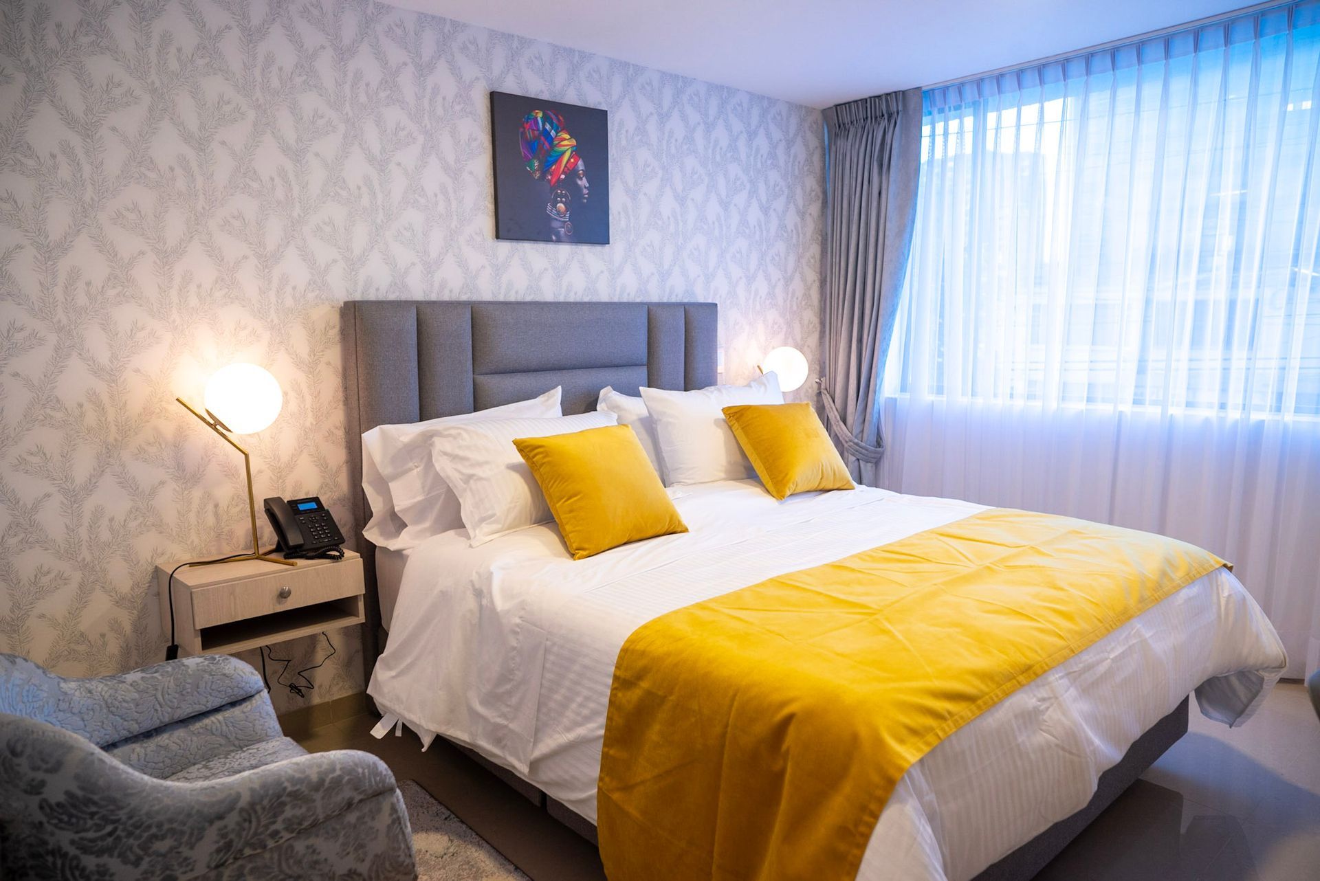 Una habitación de hotel con una cama tapizada en gris con ropa de cama blanca, almohadas decorativas amarillas y una manta amarilla.