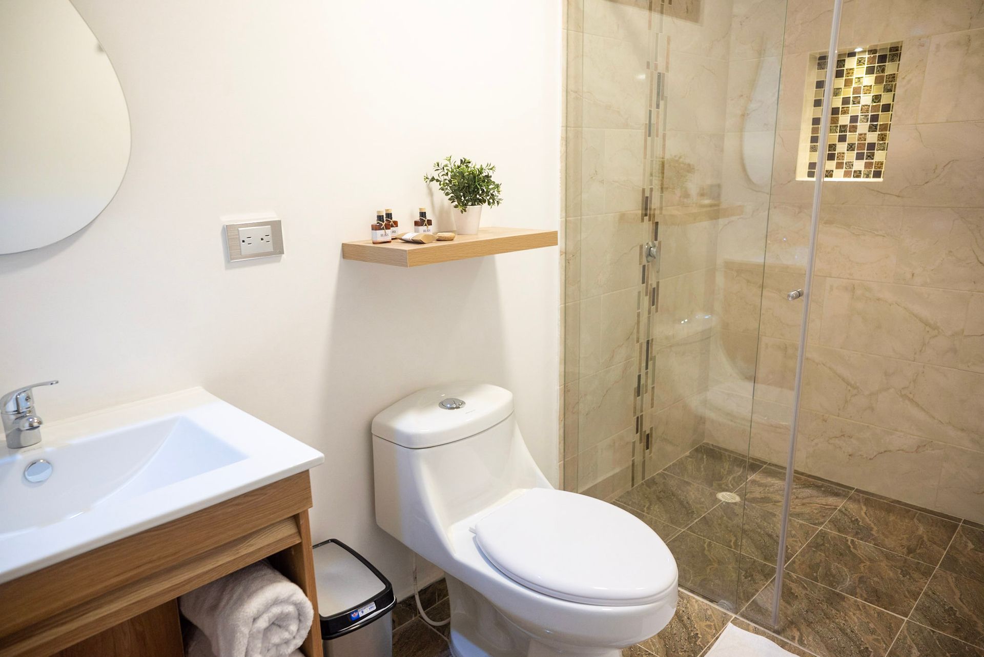 Un baño con lavabo blanco, inodoro, pequeño estante de pared con decoración y una ducha con mampara de cristal y paredes de azulejos de piedra.
