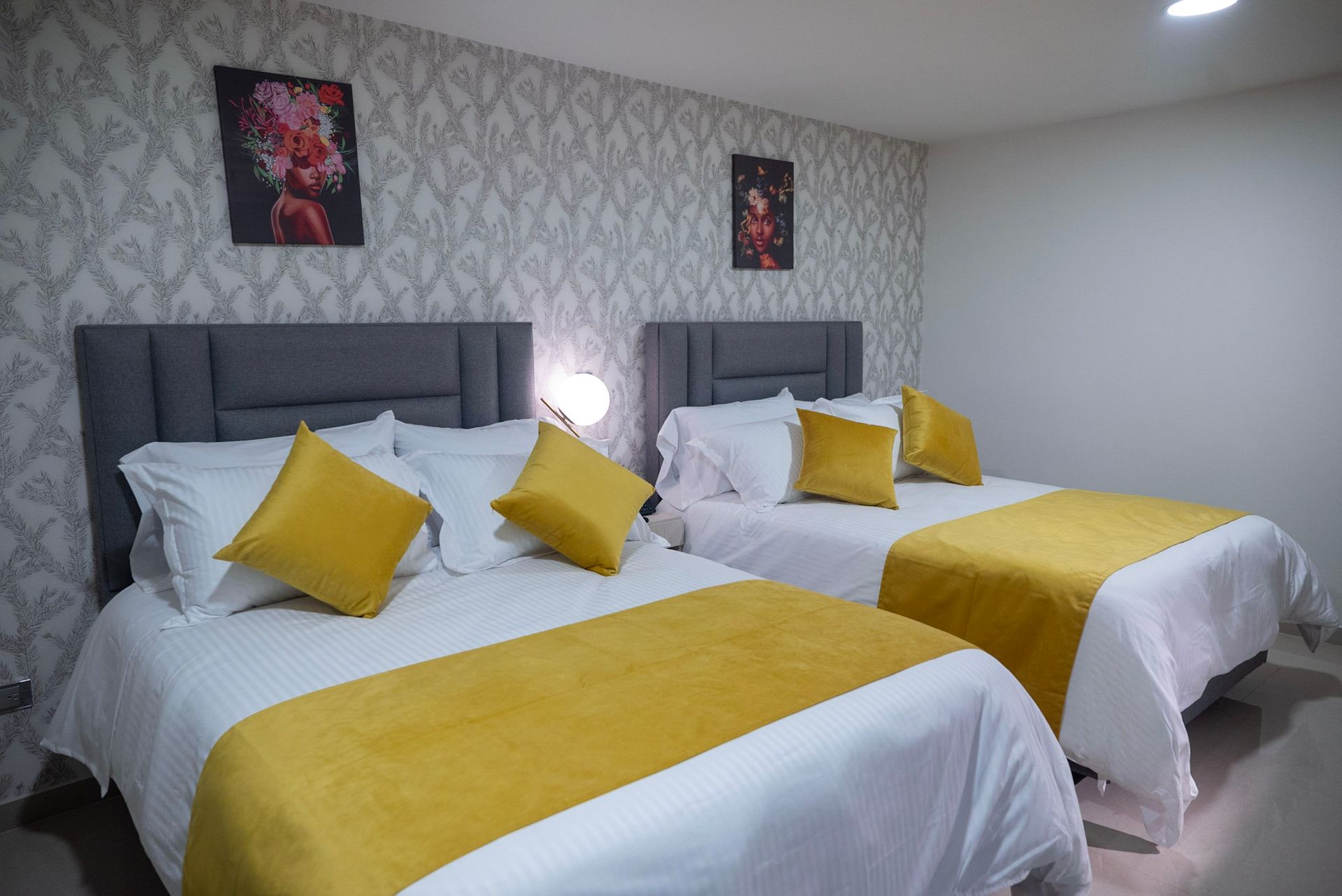 Dos camas con sábanas blancas, detalles en amarillo y cabeceras grises en una habitación con papel tapiz estampado y obras de arte enmarcadas.