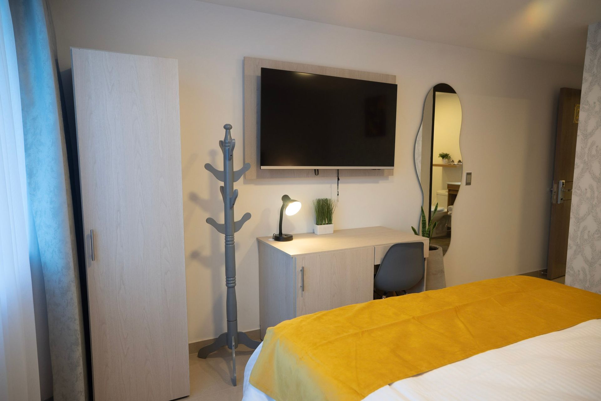 Un dormitorio con una cama con cubierta amarilla, un escritorio blanco, una silla gris, un televisor colgado, un perchero y un espejo ovalado.