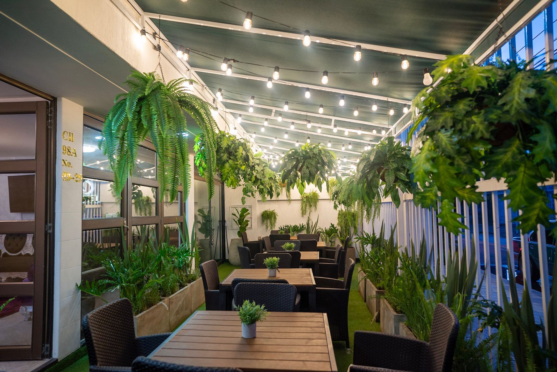 Un acogedor restaurante al aire libre con patio, mesas de madera, sillas de mimbre, helechos colgantes y cálidas luces de cadena en el techo.
