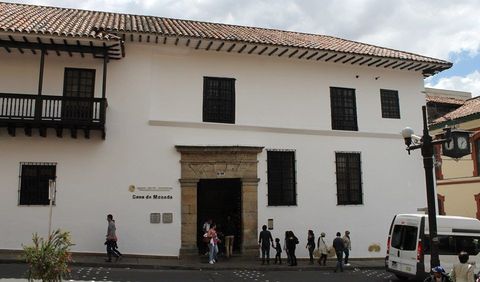 Un edificio histórico, blanco, de dos pisos, con balcones de madera oscura, una entrada con marco de piedra y gente caminando al frente.