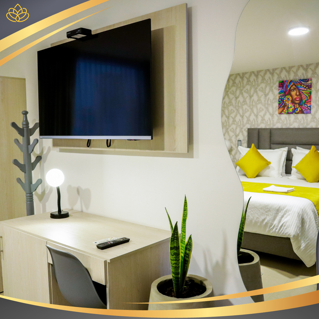 Una habitación de hotel con escritorio, televisor de pantalla plana y una planta de interior, que muestra una vista de una cama con detalles amarillos a través de un espejo.