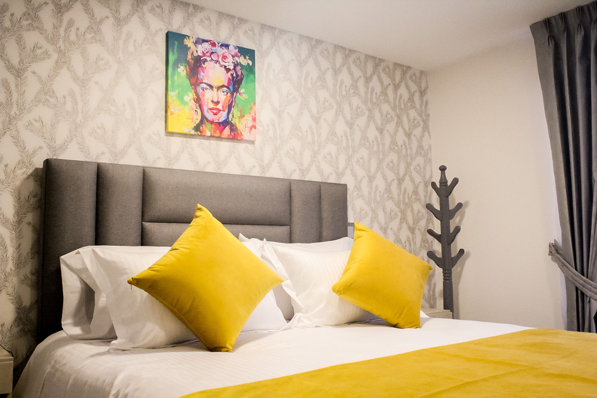 Un dormitorio con una cabecera tapizada en gris, ropa de cama blanca y dos almohadas decorativas amarillas debajo de una colorida obra de Frida Kahlo.