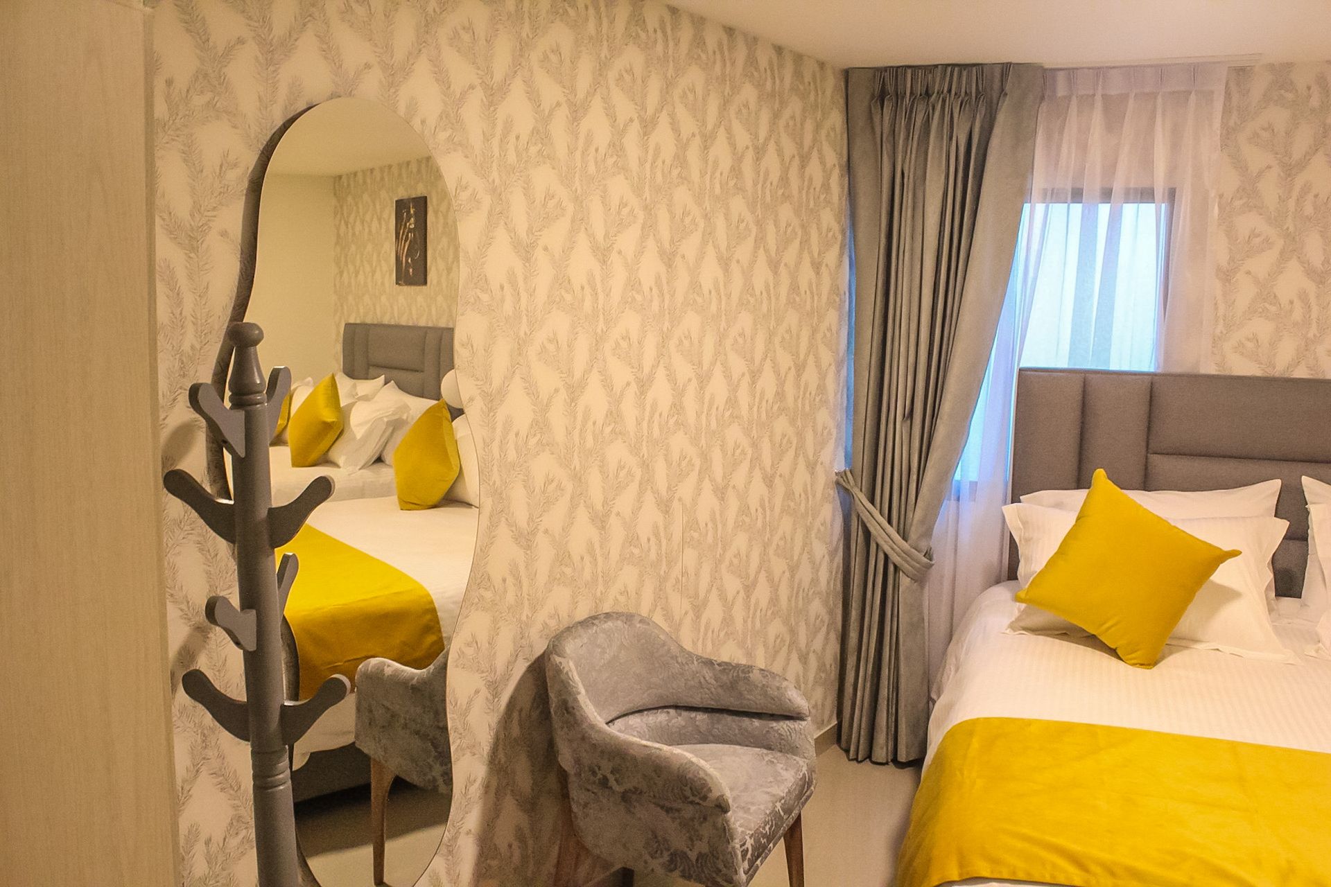 Una habitación de hotel con una cama con ropa de cama blanca y almohadas amarillas, una silla gris y un espejo arqueado que refleja la cama.