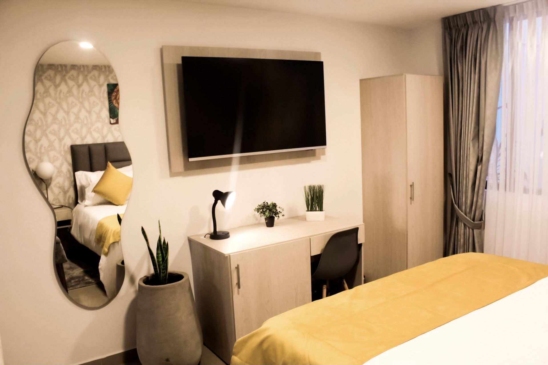 Una moderna habitación de hotel con una cama, un escritorio con lámpara y plantas, un televisor montado en la pared, un espejo alto y un armario.