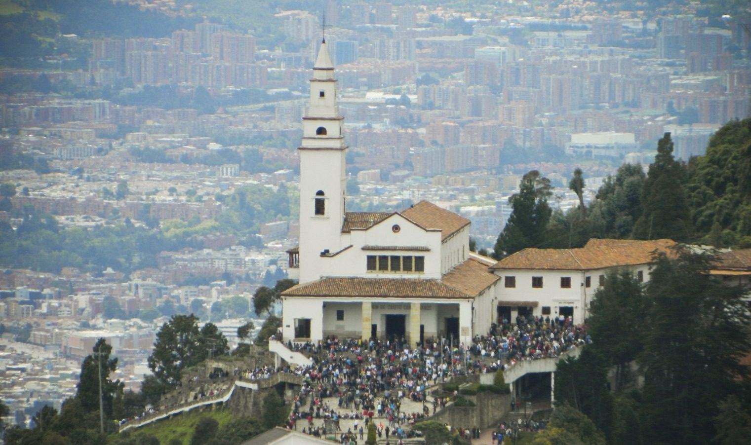 Una iglesia blanca con un alto campanario se alza sobre una montaña con vistas a una ciudad en expansión, con multitudes reunidas en su base.