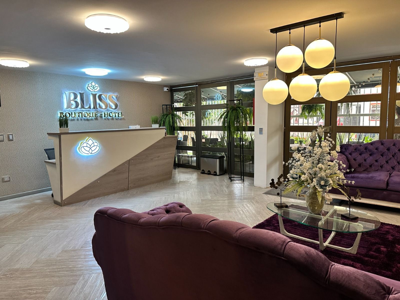 Vestíbulo del hotel con un mostrador de recepción de madera que presenta un cartel Bliss retroiluminado, sofás morados y una moderna lámpara de cristal.