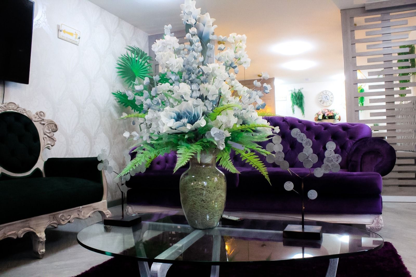 Un sofá de color morado con mechones y una silla oscura se encuentran en una habitación con un gran arreglo floral blanco sobre una mesa de café de vidrio.