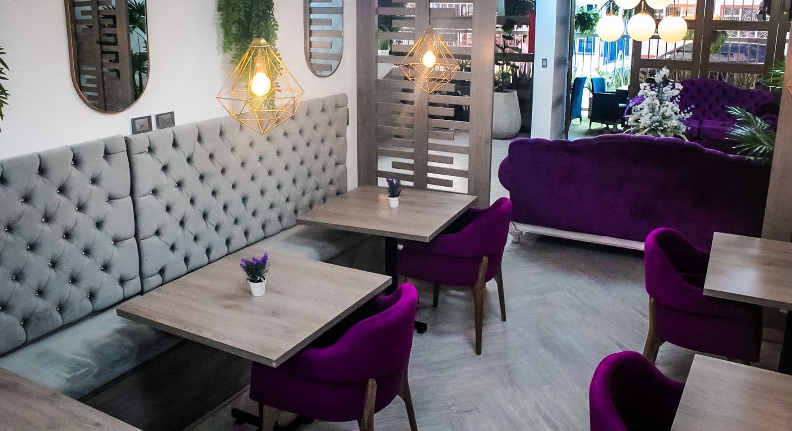 Una fotografía del interior de una elegante cafetería con cabinas con mechones grises, mesas de madera y sillas tapizadas de color violeta vibrante.