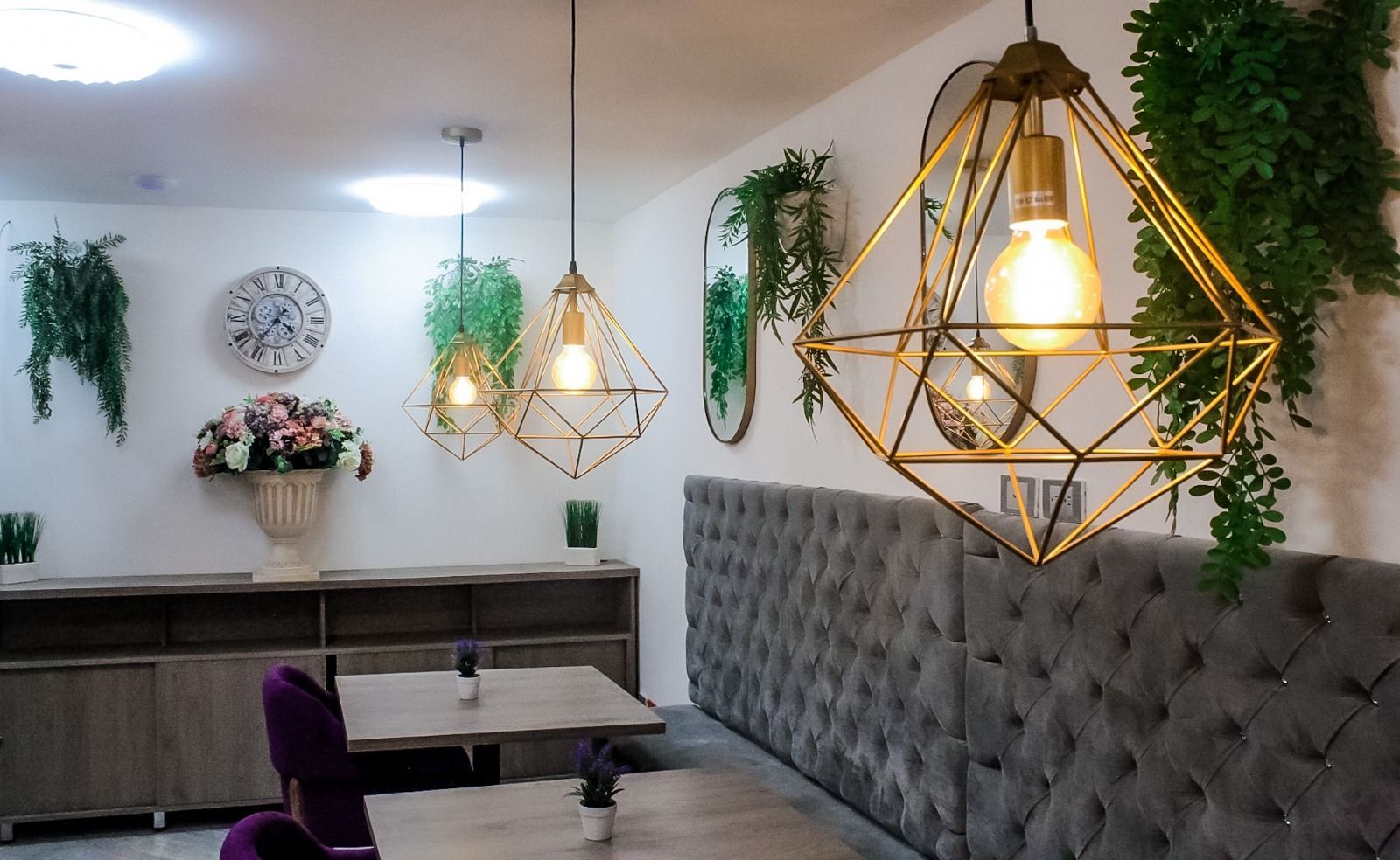 Un elegante comedor con luces colgantes geométricas doradas, una banqueta gris con mechones, mesas de madera y plantas colgantes.