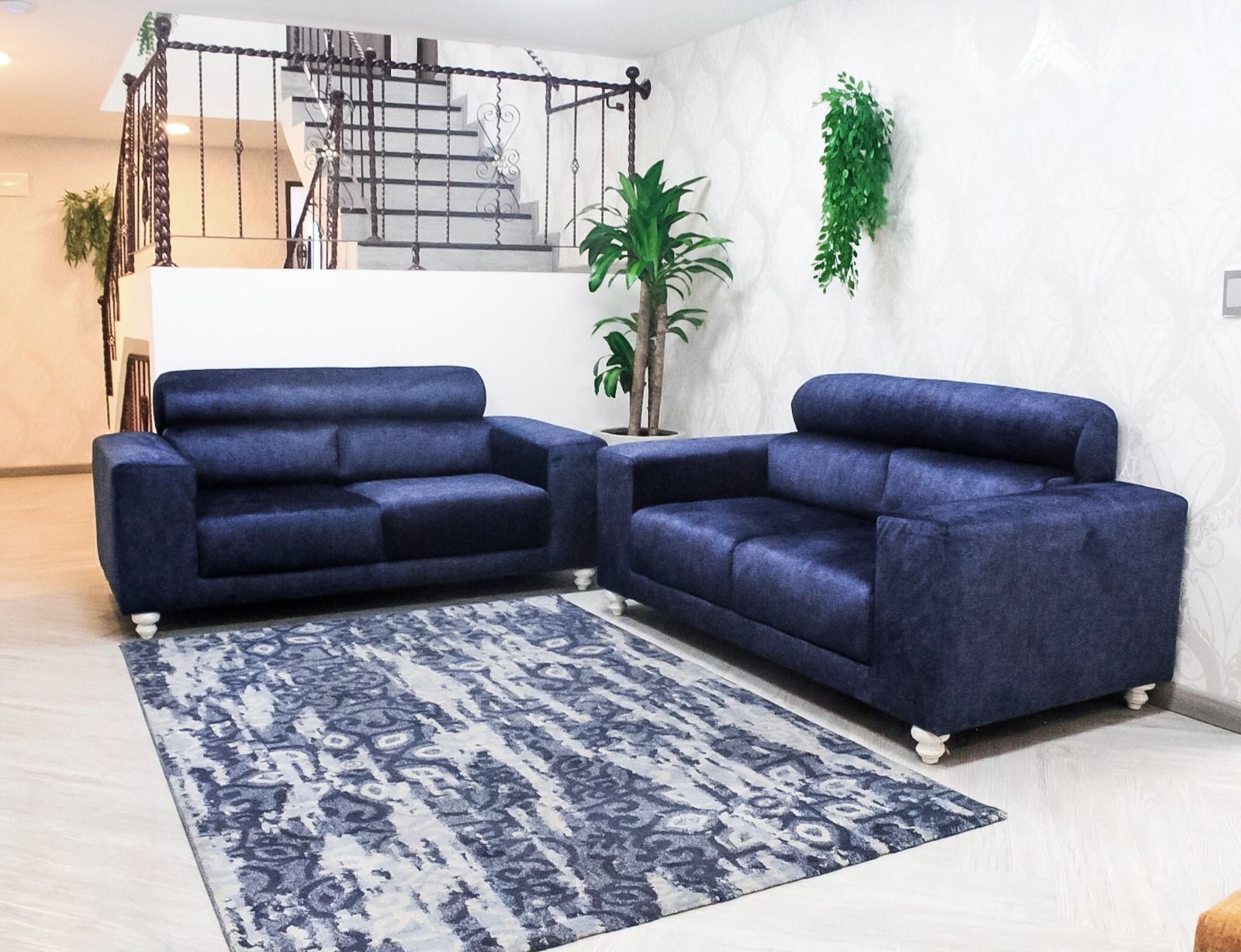 Dos sofás de tela de color azul marino dispuestos en una sala de estar con una alfombra estampada, una planta alta y una escalera.