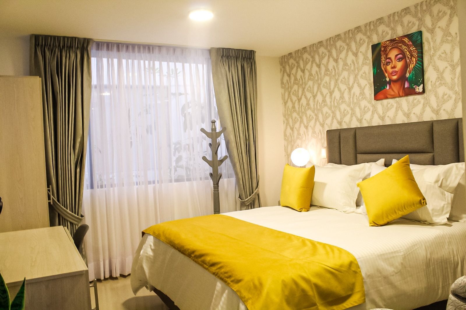 Un dormitorio con una cama con sábanas blancas y detalles en amarillo, cortinas gris claro y una pieza de arte en la pared.