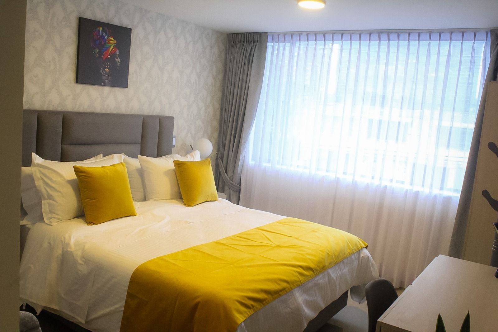 Un dormitorio con una cama con ropa de cama blanca, almohadas decorativas amarillas y una manta, y una cabecera gris sobre un papel tapiz gris.