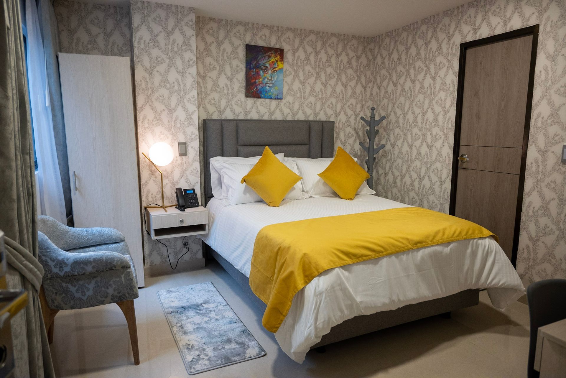 Una moderna habitación de hotel con una cama tamaño queen, almohadones y camino de mesa con detalles en color amarillo, papel tapiz estampado y un sillón azul.