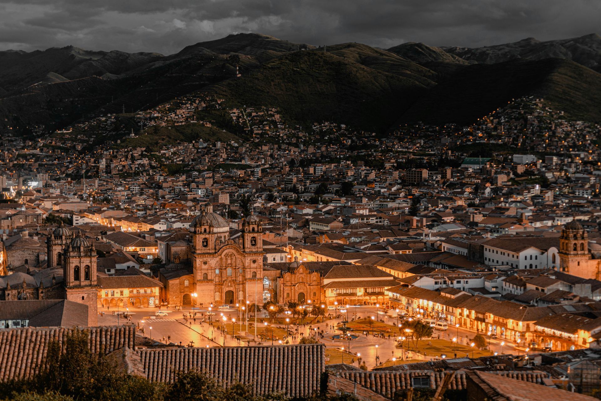 Vista nocturna de Cusco, Perú, que muestra una plaza brillantemente iluminada, edificios coloniales históricos y montañas circundantes.