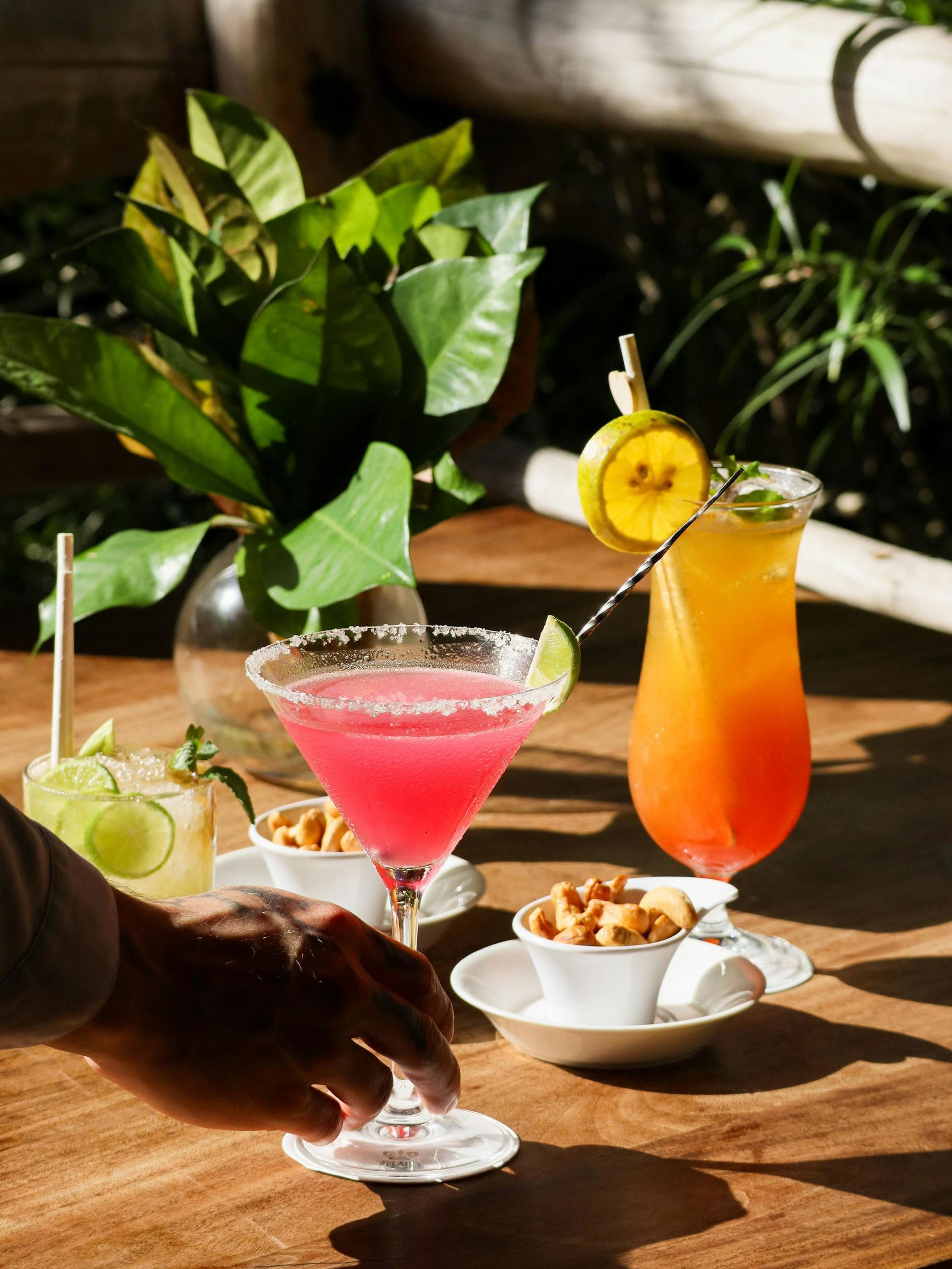 Una mano se acerca a una margarita de color rosa brillante sobre una mesa de madera junto a otros dos cócteles y pequeños tazones de nueces.