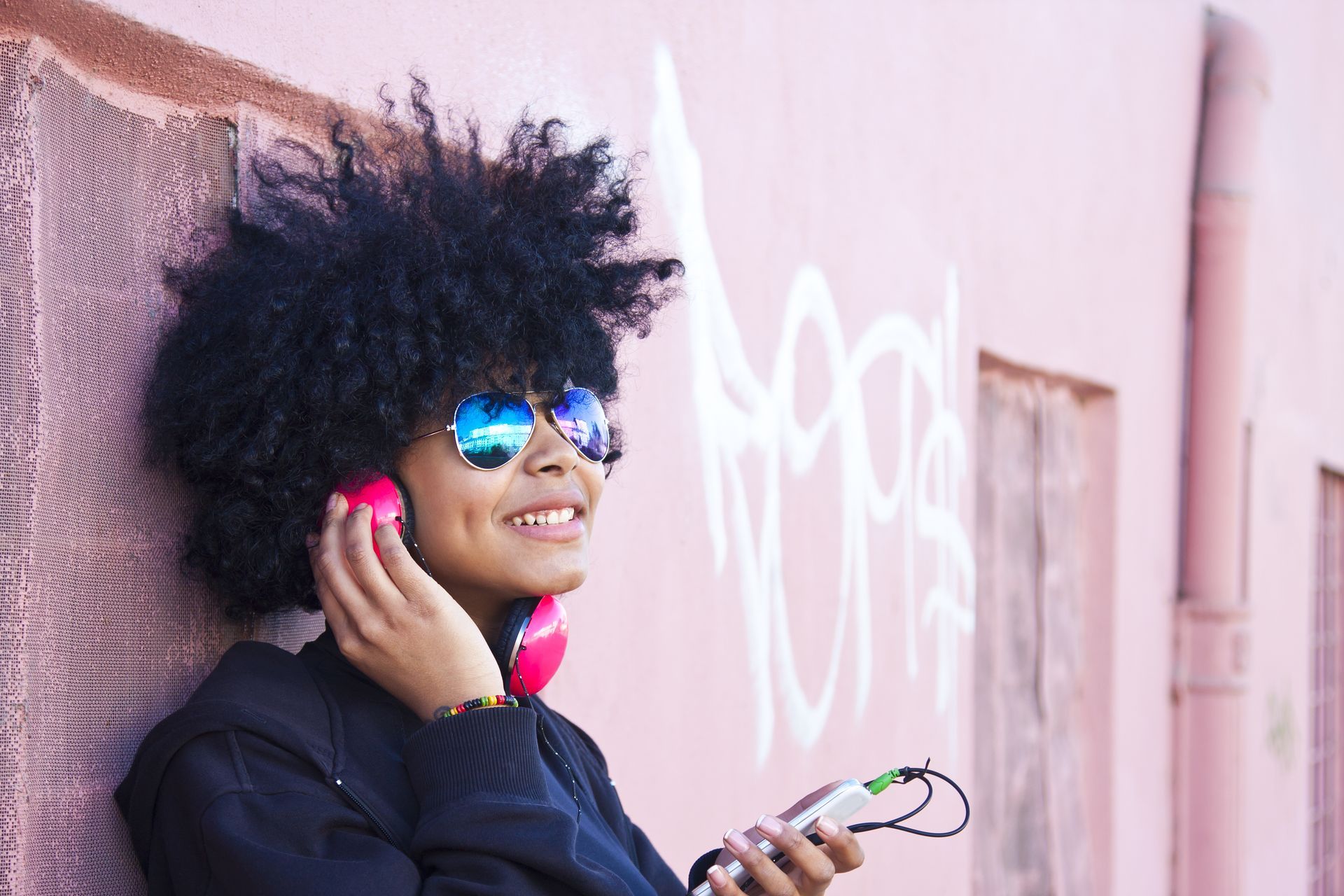 A woman wearing headphones and sunglasses is leaning against a pink wall.