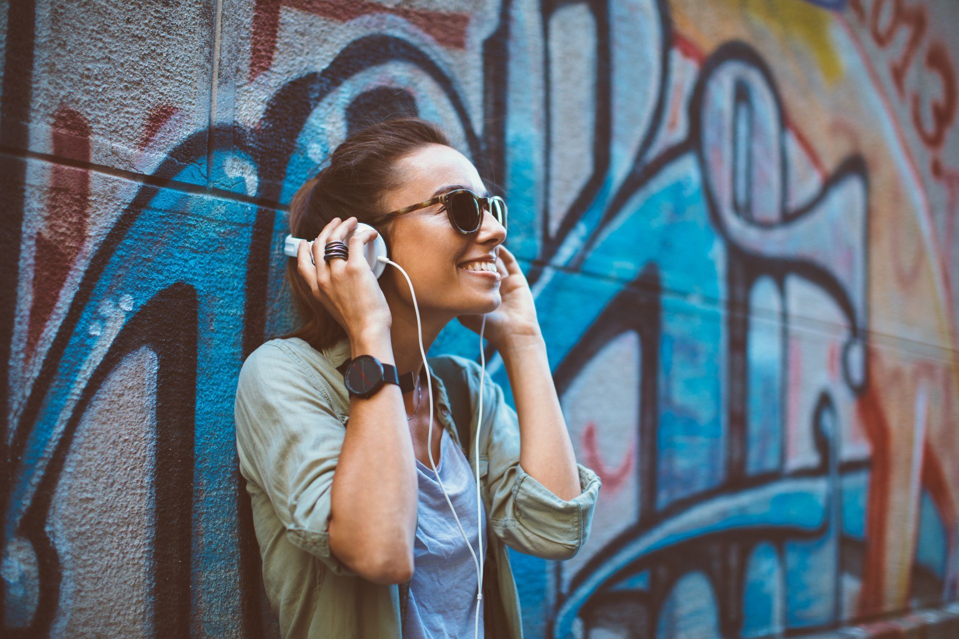 A woman is listening to music with headphones in front of a graffiti wall.
