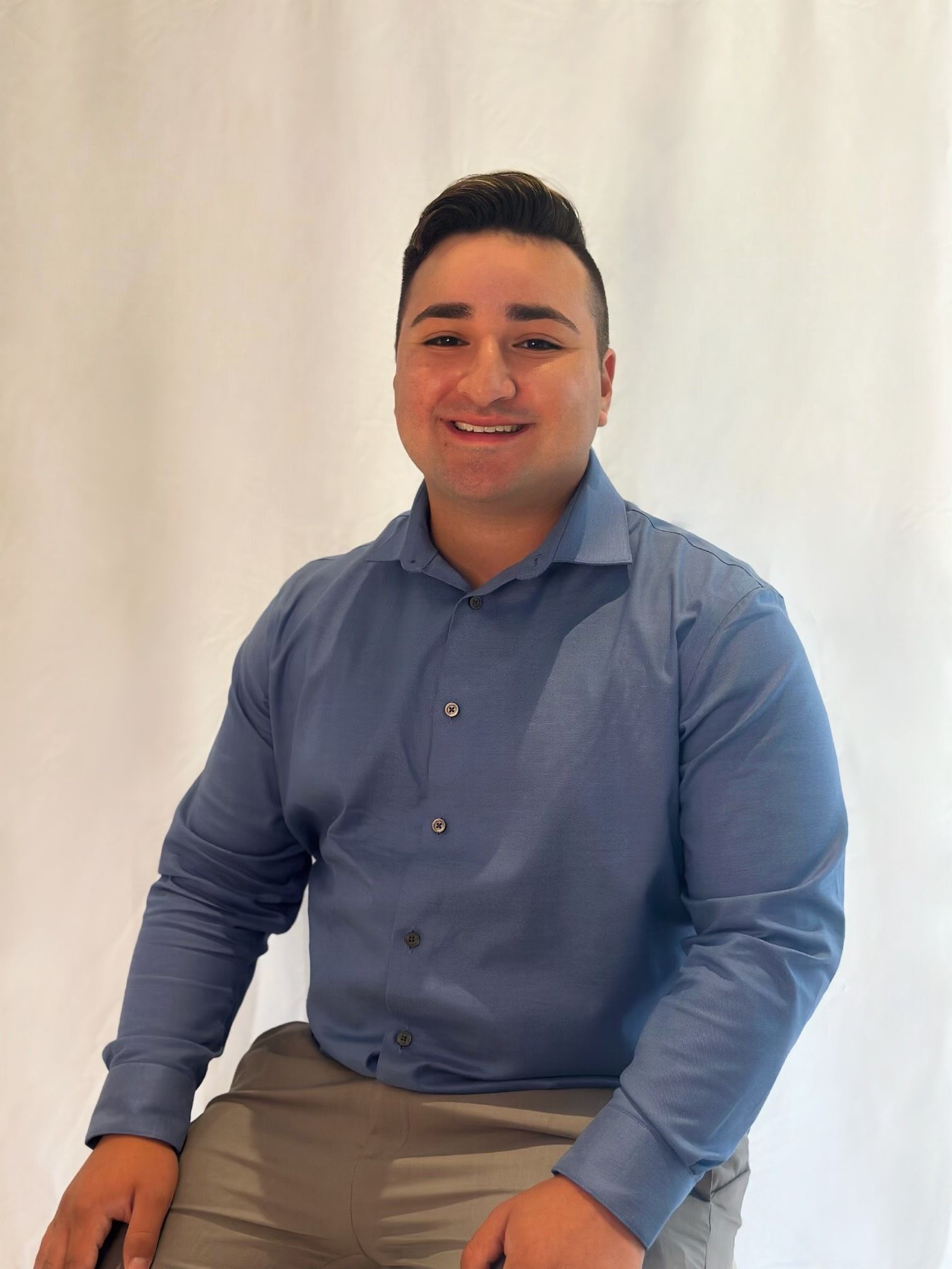 A man in a blue shirt is sitting on a stool and smiling.