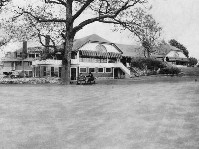 Black and white photo of a large building with a covered porch and lawn. Two figures are seated near a tree.