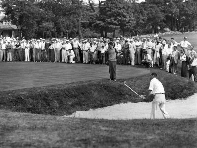 Golfer playing from sand trap with large crowd watching behind him.