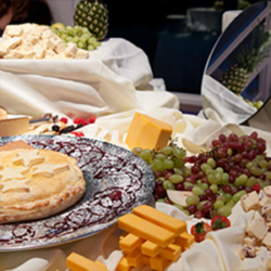 Buffet spread: cheeses, grapes, pastry, pineapple, and other foods on a draped table with a decorative mirror.